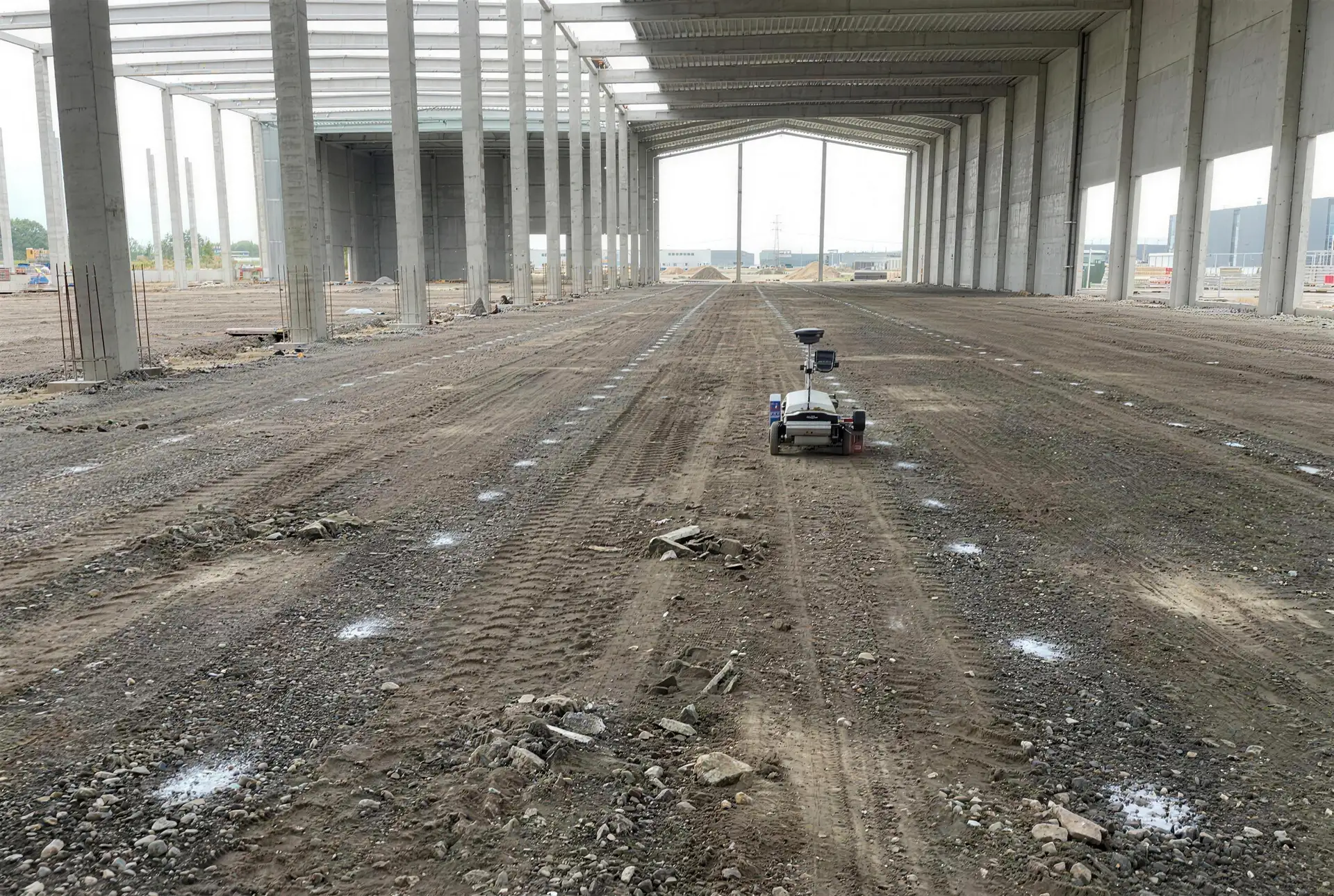 Hal in aanbouw met betonkolommen; rijen witte uitzetstippen markeren de indeling op de ondergrond en onze robotplotter rijdt de volgende rij